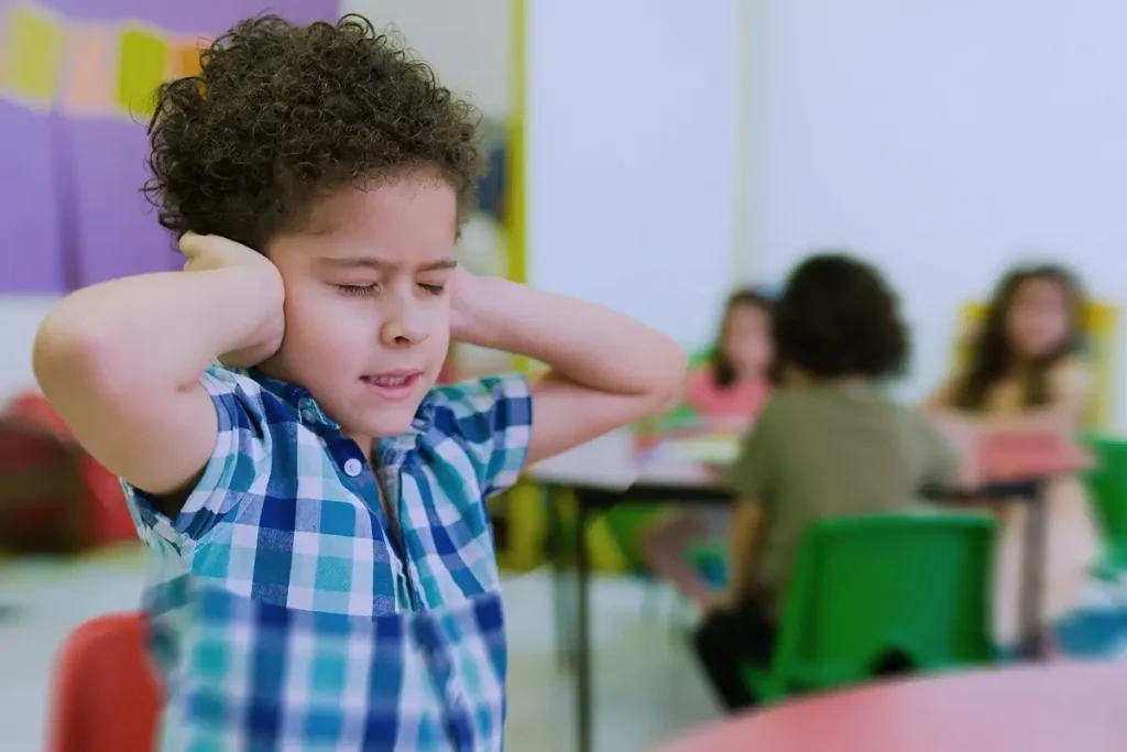 Bambino in classe con le mani sulle orecchie, esprimendo disagio sensoriale, mentre altri bambini sono seduti ai tavoli sullo sfondo.