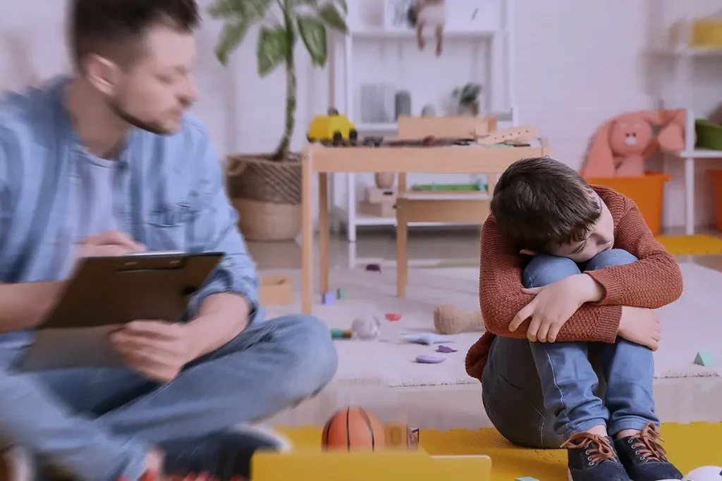 Bambino seduto a terra con le ginocchia abbracciate, mentre uno specialista prende appunti osservandolo in un ambiente giocoso.