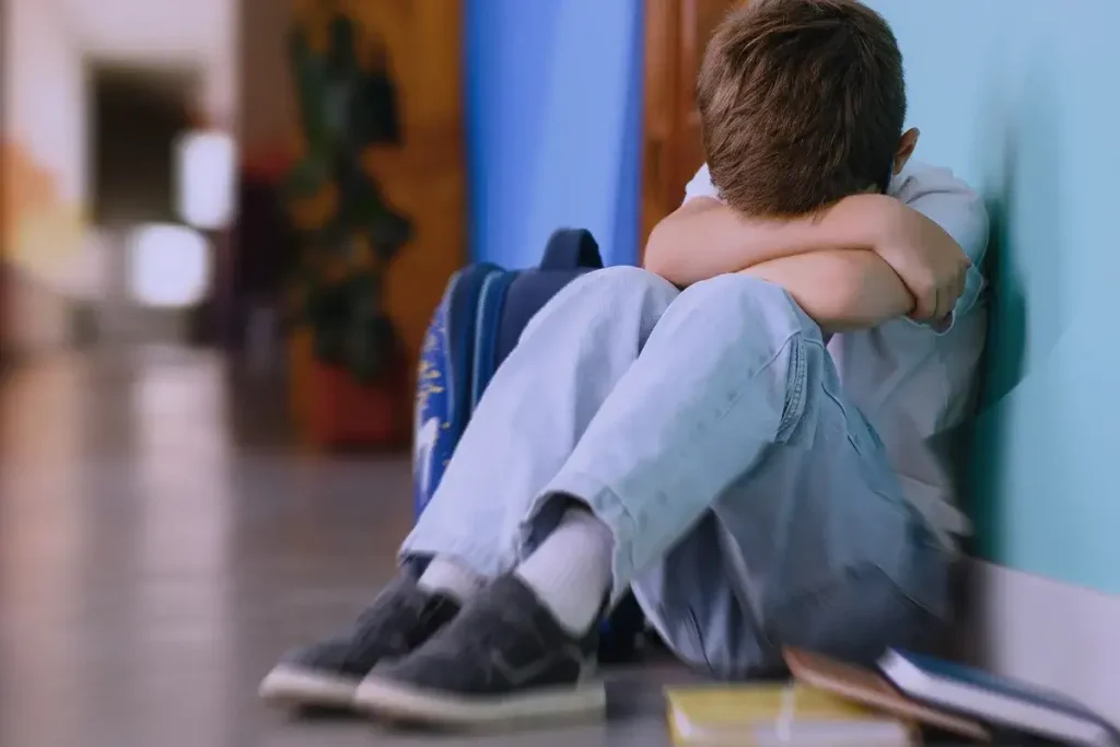 Bambino seduto a terra in un corridoio scolastico, con la testa appoggiata sulle braccia e libri sparsi accanto a lui.