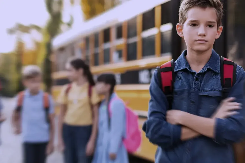 Ragazzo con espressione triste e braccia incrociate davanti a uno scuolabus, mentre un gruppo di coetanei parla tra loro in secondo piano.