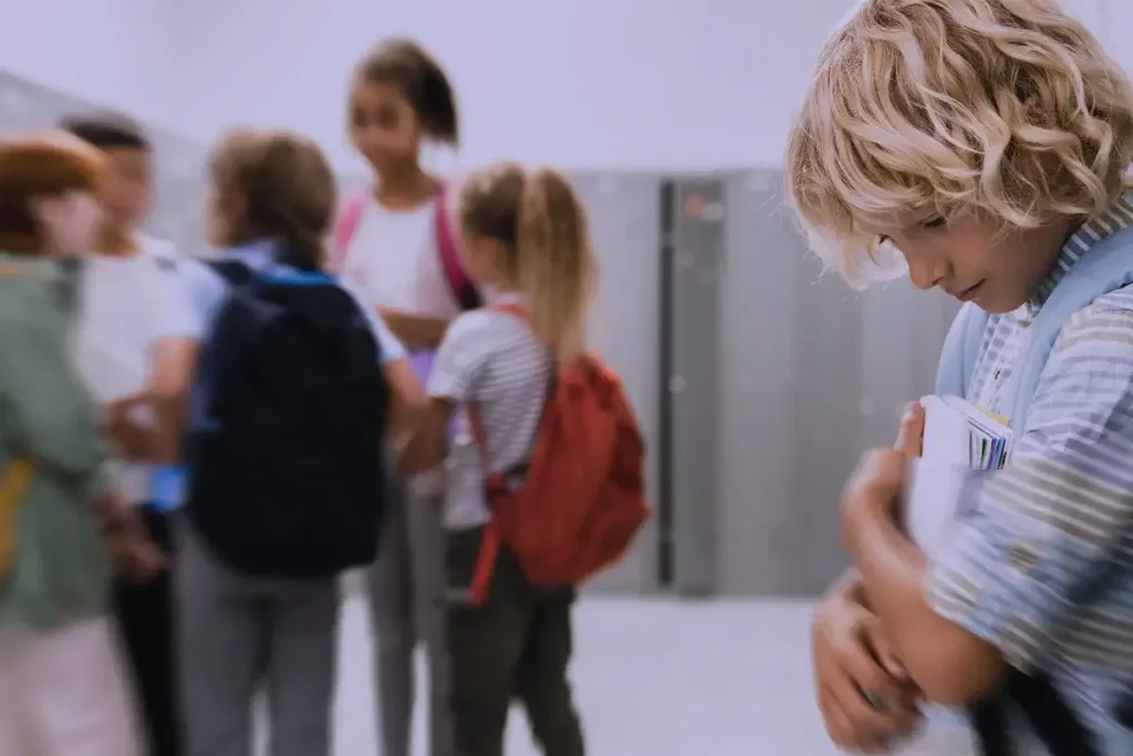 Bambino triste appoggiato agli armadietti della scuola, stringe un libro mentre un gruppo di compagni parla tra loro in lontananza.