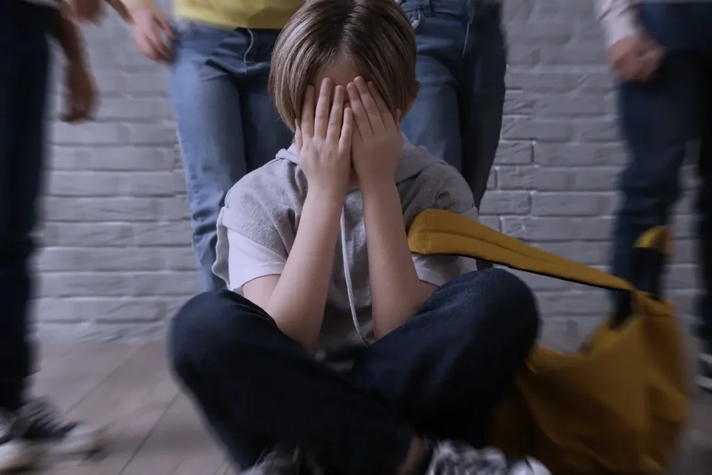 Bambino seduto a terra con il viso coperto dalle mani, circondato da coetanei che lo deridono e lo indicano.