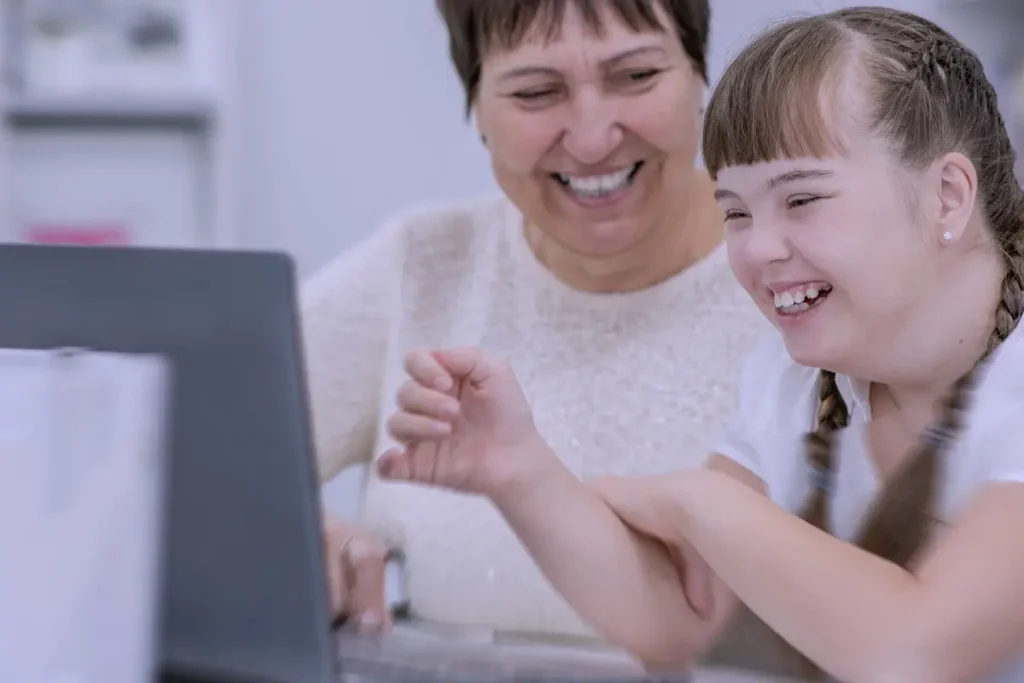 Bambina con sindrome di Down che sorride mentre utilizza un computer portatile con una donna adulta, entrambe felici in un ambiente scolastico o biblioteca.